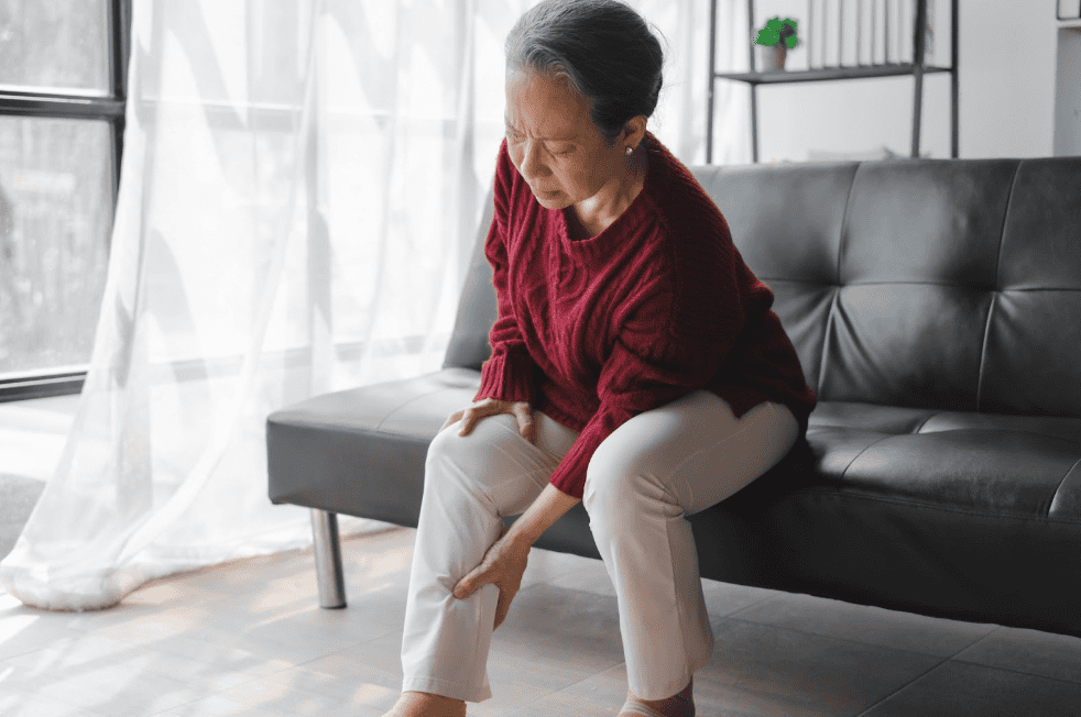 Elderly Asian woman sitting on couch grabbing her leg because of sciatica pain